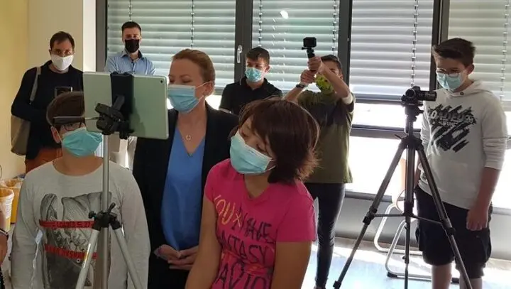 Schülerinnen und Schüler mit Masken vor Tablets und Kameras auf Stativen, Erwachsene schauen zu