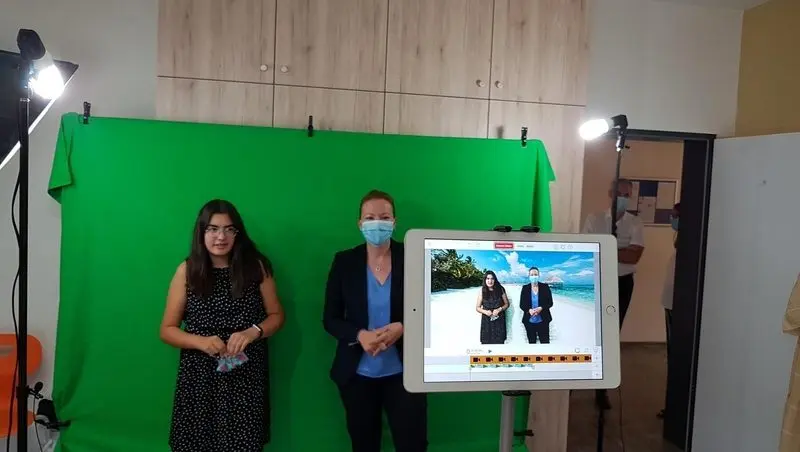 Schülerin und Besucherin vor Greenscreen, Tablet zeigt beide vor Strandkulisse