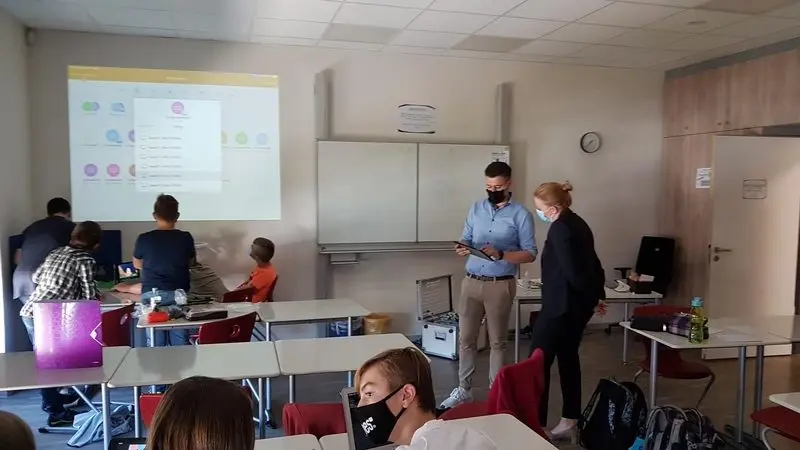Besucherin und Lehrkraft mit Tablet im Klassenraum, Beamer zeigt eine App-Übersicht