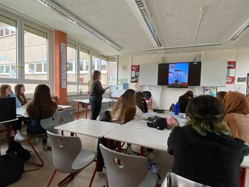 Schülerinnen im Klassenraum schauen auf einen Bildschirm mit britischer Flagge, eine Schülerin steht vorne