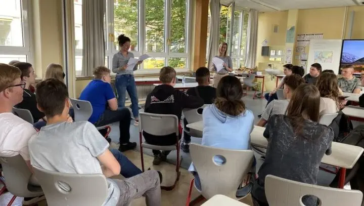 Zwei Frauen lesen vor einer Klasse aus Blättern vor, die Schülerinnen und Schüler sitzen im Halbkreis
