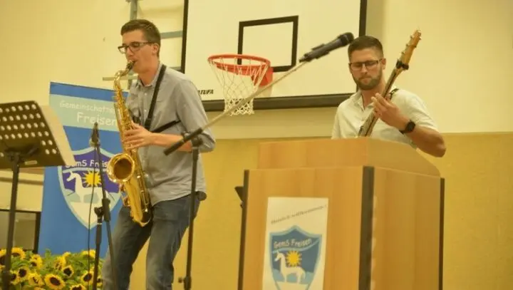 Zwei Musiker mit Saxofon und E-Bass auf der Bühne der Turnhalle neben einem Rednerpult mit Schullogo