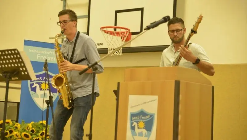 Zwei Musiker mit Saxofon und E-Bass auf der Bühne der Turnhalle neben einem Rednerpult mit Schullogo