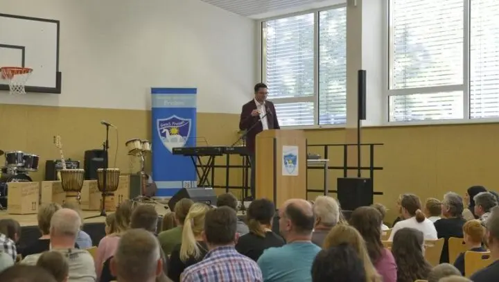 Redner am Pult in der Sporthalle vor Publikum, daneben Musikinstrumente und Schulbanner
