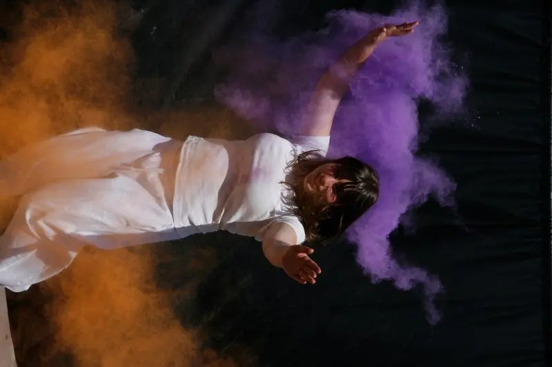 Fotografie: Person in weißem Kleid zwischen orangen und violetten Farbpulverwolken vor schwarzem Hintergrund