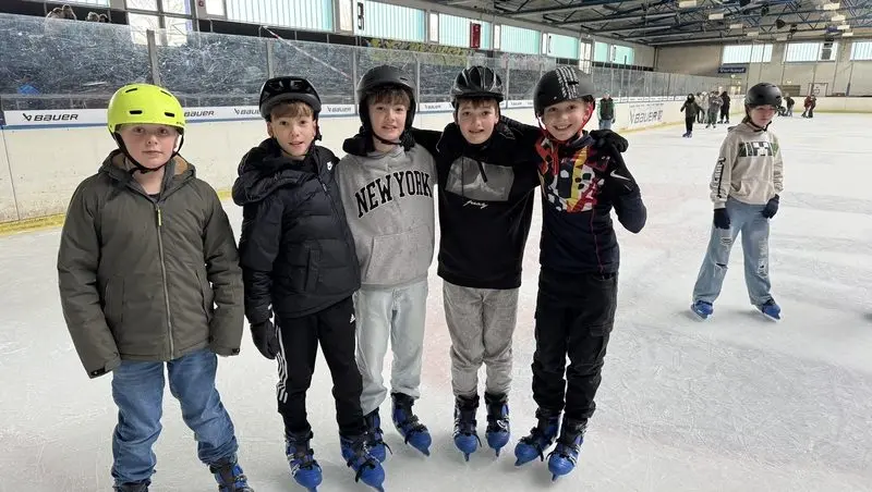 Fünf Kinder mit Helmen und Schlittschuhen stehen Arm in Arm auf der Eisfläche einer Eishalle