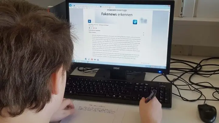 Schüler sitzt vor einem Bildschirm mit der Webseite „Fakenews erkennen“ und macht Notizen