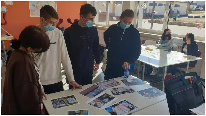 Jugendliche mit Masken betrachten ausgedruckte Fotos auf einem Tisch im Klassenraum