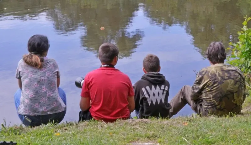Vier Jugendliche sitzen von hinten gesehen am Ufer eines Teichs, eine Kamera dabei