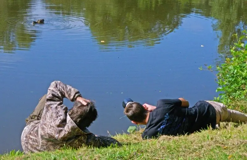 Zwei Personen liegen am Teichufer und fotografieren eine Ente auf dem Wasser