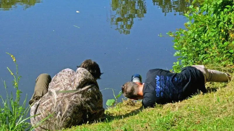 Zwei Personen liegen am grasigen Ufer und fotografieren übers Wasser, eine in Tarnkleidung