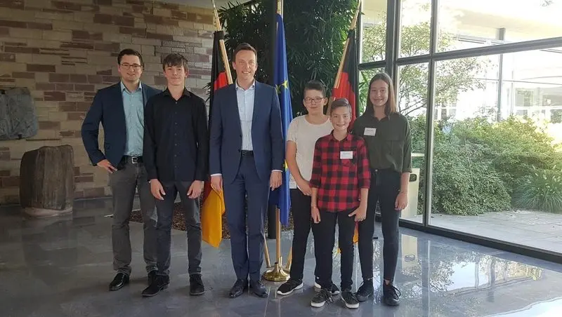 Gruppenfoto: zwei Männer im Sakko und vier Jugendliche vor Deutschland- und Europaflaggen in einem Foyer