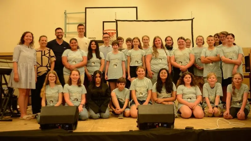 Gruppenfoto der Klasse in einheitlichen grauen T-Shirts mit Erwachsenen auf der Bühne der Turnhalle