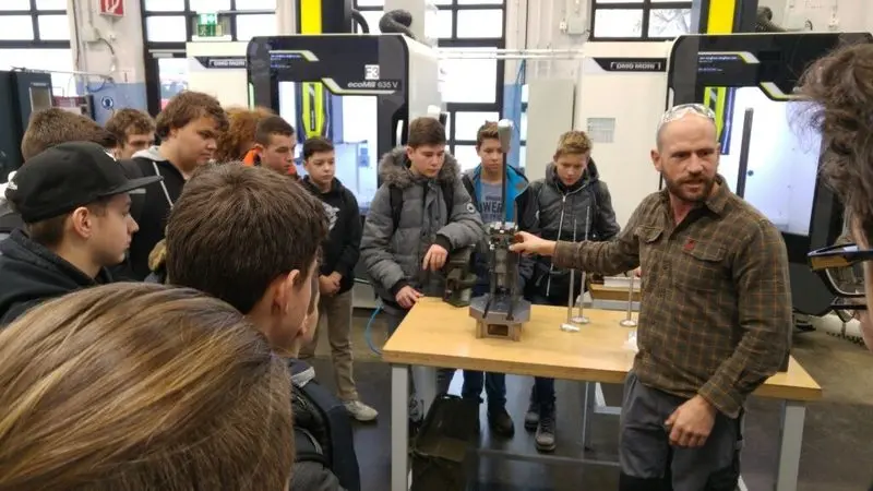 Ausbilder erklärt Jugendlichen ein Werkstück an einem Tisch vor CNC-Maschinen