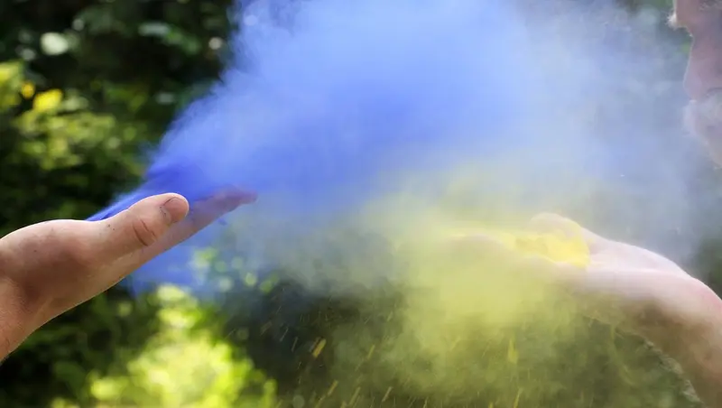 Blaues und gelbes Farbpulver wird von zwei Händen in die Luft gepustet