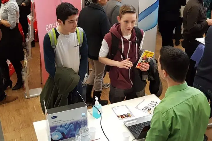 Zwei Jugendliche sprechen an einem Messestand mit einem Mann, einer hält eine Broschüre