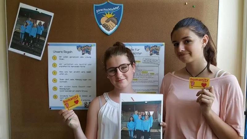 Zwei Schülerinnen mit Humanity Cards und einem Foto vor der Pinnwand der Streitschlichter