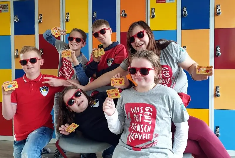 Sechs Kinder und Jugendliche mit roten Sonnenbrillen halten „humanity“-Karten vor bunten Spinden