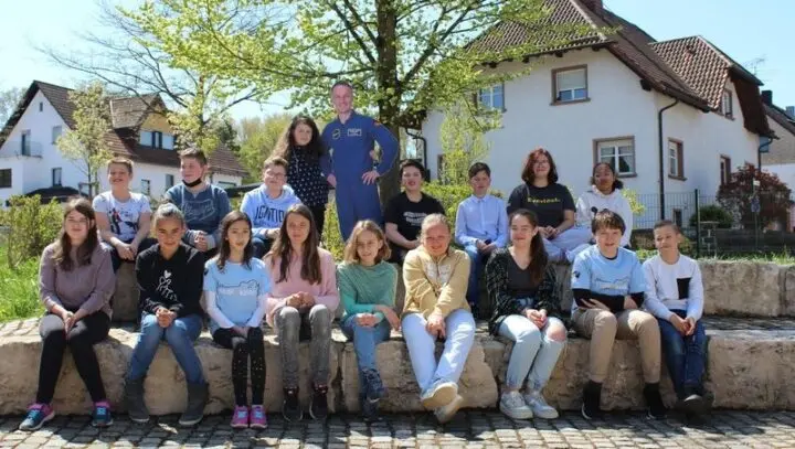 Schulklasse sitzt auf einer Steinmauer im Freien, in der Mitte der Aufsteller eines Astronauten