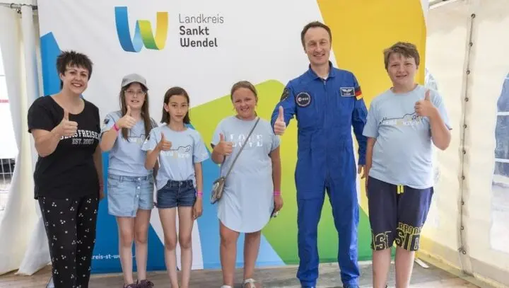 Vier Mädchen, ein Junge, eine Frau und ein Astronaut im Raumanzug zeigen Daumen hoch