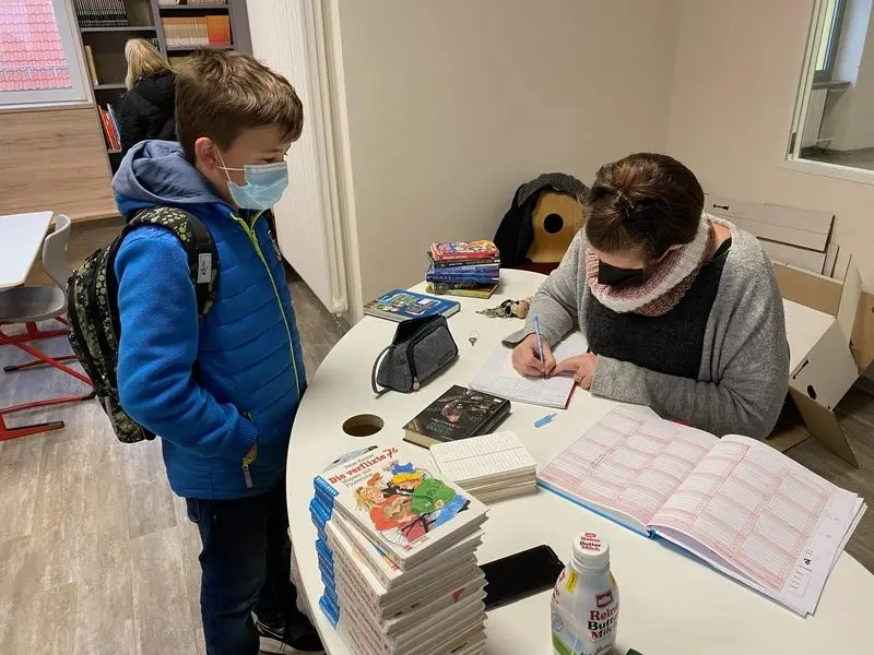 Schüler mit Maske leiht ein Buch aus, eine Frau trägt es am Tisch in eine Liste ein