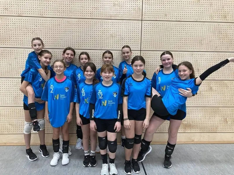 Größere Mädchen-Volleyballmannschaft in blauen Trikots, zwei Spielerinnen werden von Mitspielerinnen getragen