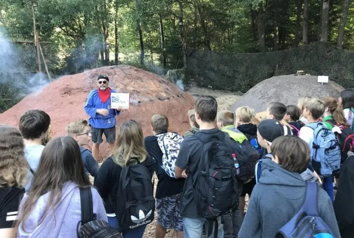 Mann mit rotem Halstuch erklärt einer Schülergruppe vor einem rauchenden Kohlemeiler im Wald eine Schautafel