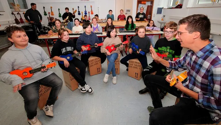 Schülerinnen und Schüler mit bunten Ukulelen sitzen auf Kartons, vorn eine erwachsene Person mit Ukulele