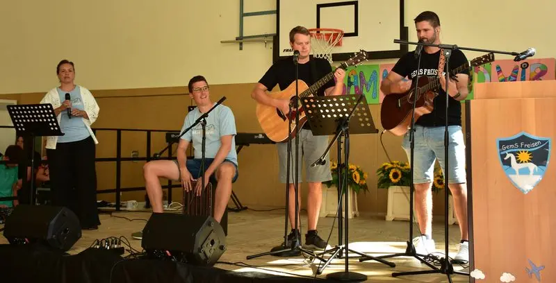 Lehrerband mit zwei Gitarren, Cajón und Sängerin auf der Bühne der Turnhalle