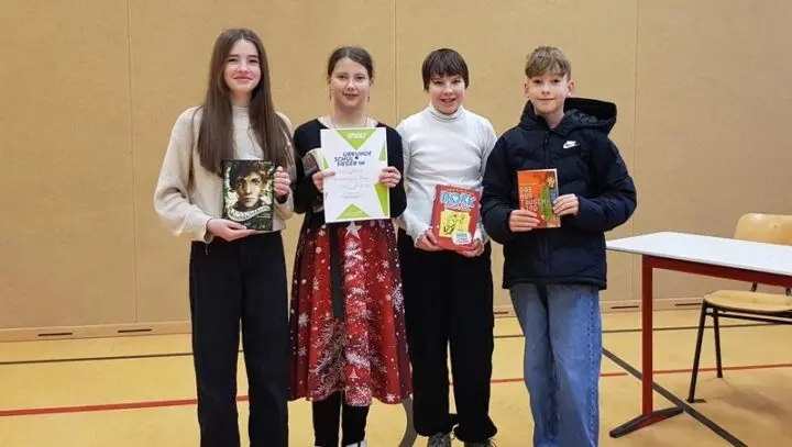Vier Schülerinnen und Schüler mit Büchern, eine hält eine Urkunde „Schulsieger:in“