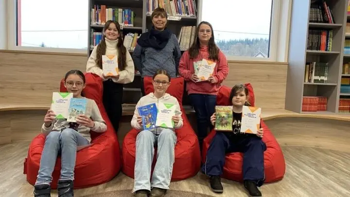 Fünf Schülerinnen mit Urkunden und Büchern, teils auf roten Sitzsäcken, in der Bibliothek, dahinter eine Frau