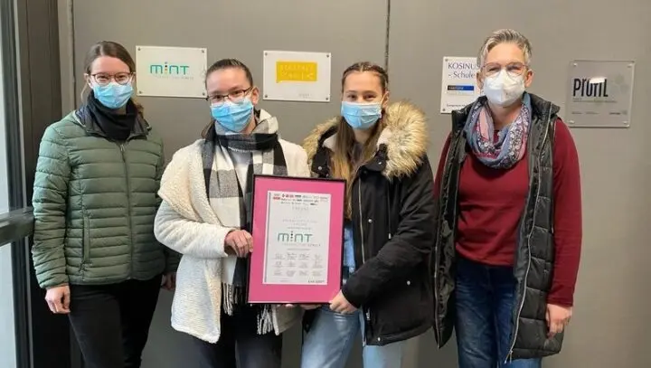 Vier Personen mit Masken halten die gerahmte Urkunde „MINT-freundliche Schule“ vor einer Wand mit Schulplaketten