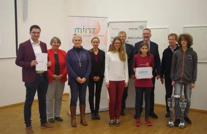 Gruppe mit MINT-Urkunde und -Schild vor Aufstellern, vorne zwei Roboter