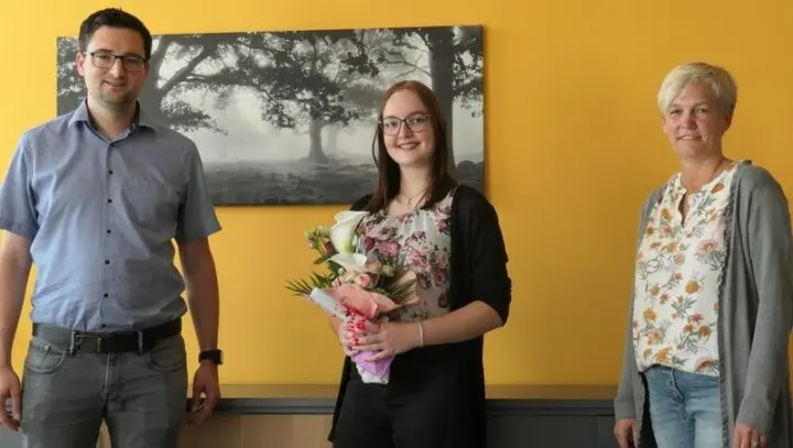 Eine Person mit Blumenstrauß steht zwischen einem Mann und einer Frau vor einer gelben Wand