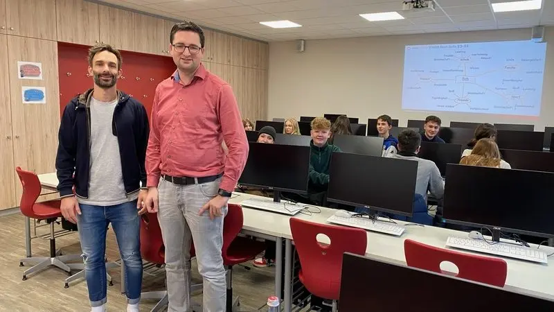 Zwei Männer stehen im Computerraum, dahinter Jugendliche und eine Mindmap am Beamer