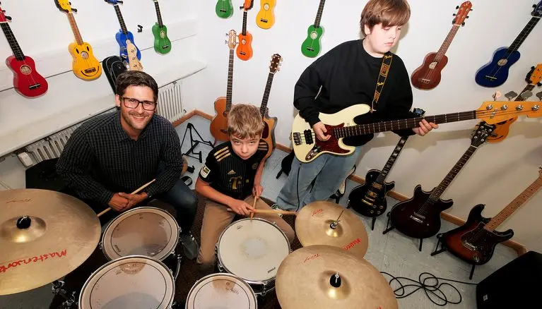 Musikraum: ein Erwachsener und zwei Schüler mit Schlagzeug und Gitarre, an der Wand hängen Ukulelen und Gitarren