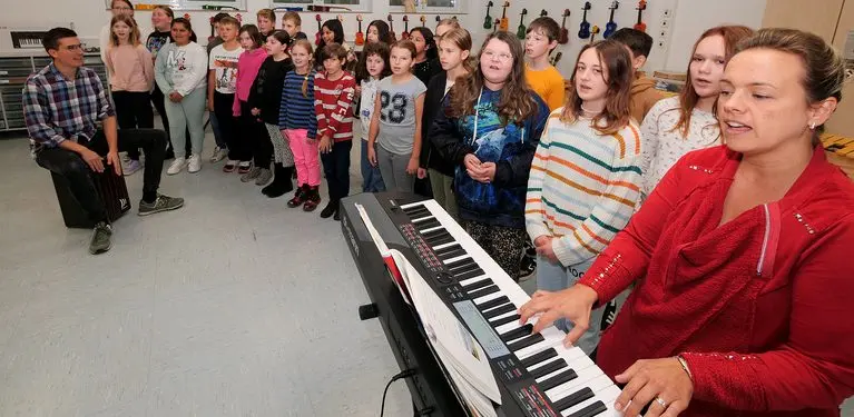Eine Person spielt Keyboard, dahinter steht eine Gruppe Schülerinnen und Schüler im Musikraum