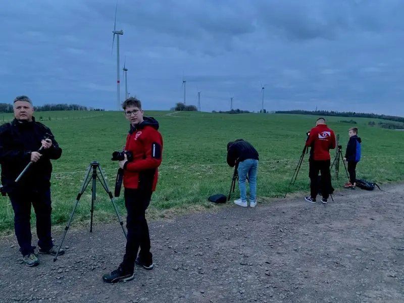 Fünf Personen mit Kameras und Stativen auf einem Feldweg in der Dämmerung, im Hintergrund Windräder