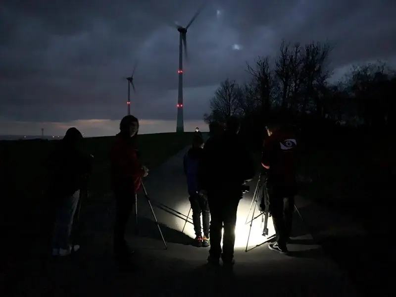 Gruppe mit Stativen fotografiert nachts auf einem Feldweg, im Hintergrund beleuchtete Windräder