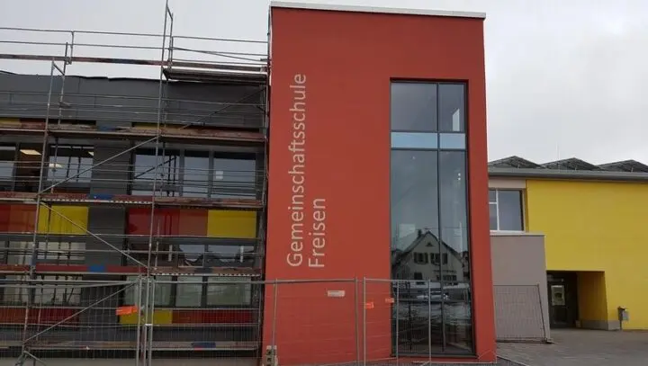 Rot verputzter Gebäudeteil mit Schriftzug „Gemeinschaftsschule Freisen“, daneben eingerüsteter Neubau