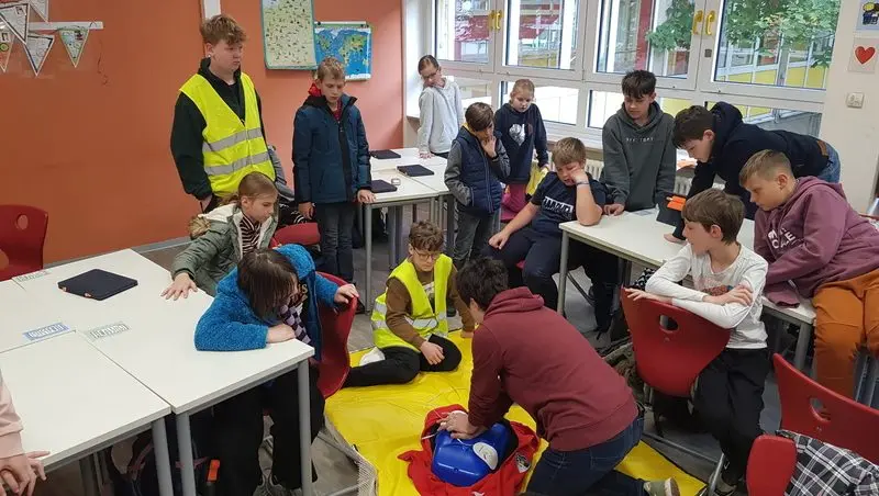 Schülerinnen und Schüler üben im Klassenraum einen Notfall, eine Person kniet auf einer gelben Decke an der Notfallausrüstung