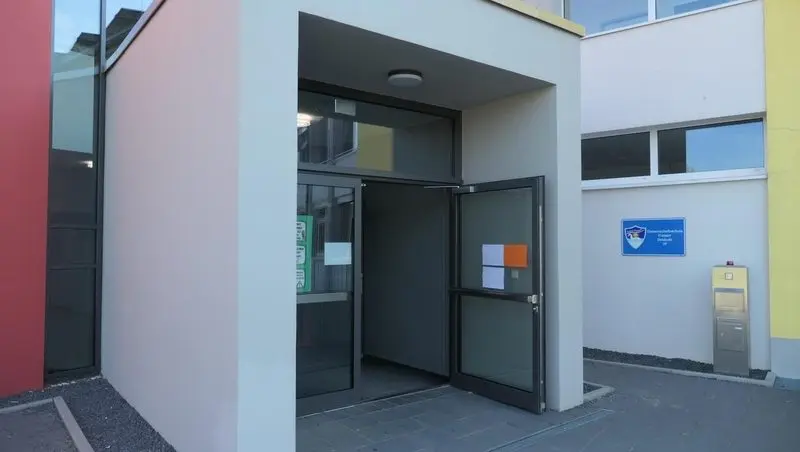 Geöffnete Eingangstür des Schulgebäudes mit Aushängen und Schulschild daneben
