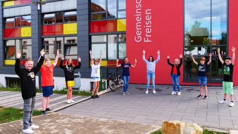 Neun Kinder stehen mit Abstand und erhobenen Armen vor dem Gebäude der Gemeinschaftsschule Freisen