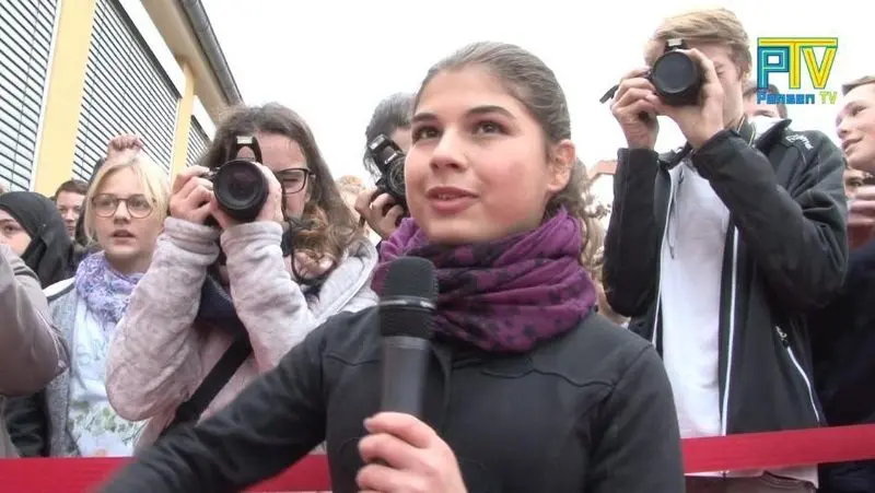 Jugendliche spricht ins Mikrofon, dahinter Personen mit Kameras, Logo „PTV Pensen TV“