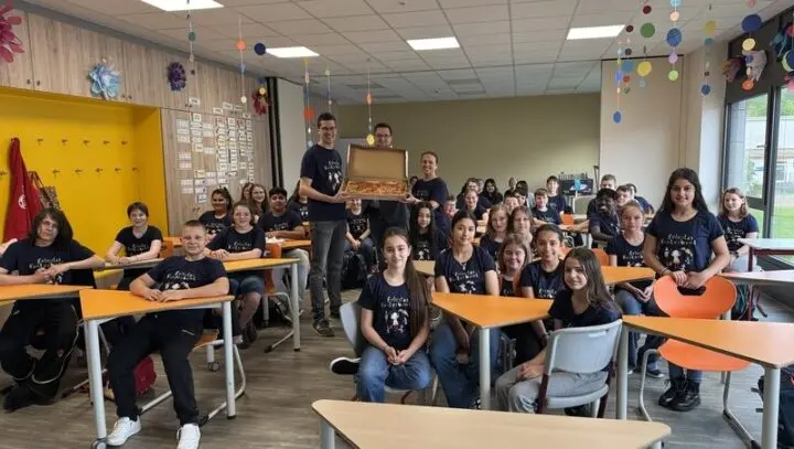 Klasse in „Felicitas Kunterbunt“-T-Shirts im Klassenraum, zwei Erwachsene halten einen Pizzakarton
