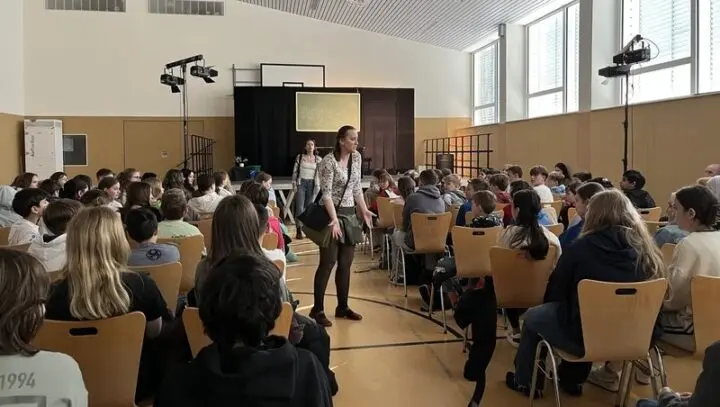 Schauspielerin spricht in der Turnhalle vor Schülerinnen und Schülern, die auf Stühlen sitzen
