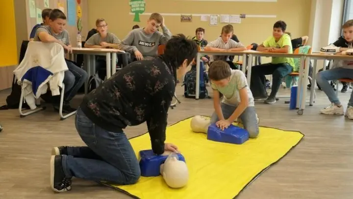 Ausbilderin und Schüler üben Herzdruckmassage an Übungspuppen, die Klasse schaut zu