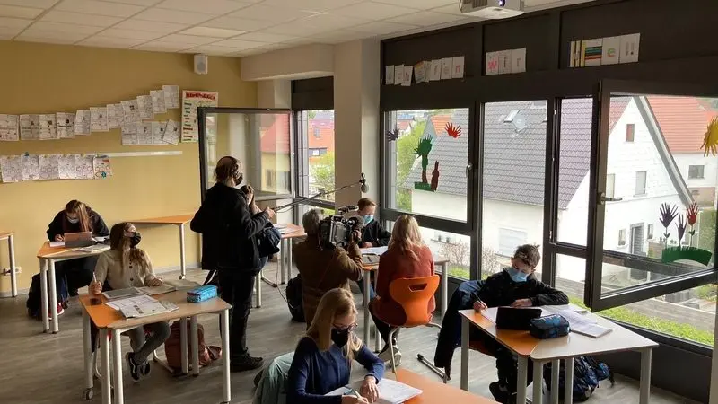 Kamerateam filmt in einem hellen Klassenraum mit großen Fenstern, Jugendliche arbeiten an ihren Tischen