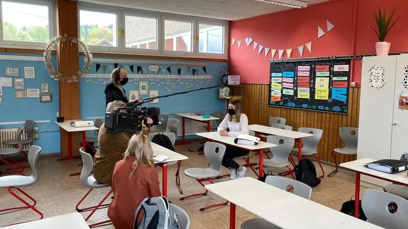 Fernsehteam filmt ein Interview mit einer Schülerin mit Maske an einem Tisch im Klassenraum