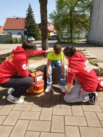 Zwei Schulsanitäter in roten Jacken mit Notfallrucksack kümmern sich um eine sitzende Person